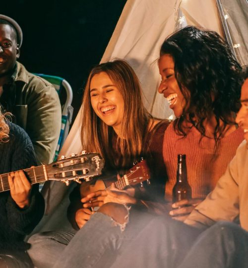 我们的服务 A diverse group of friends playing guitar and ukulele by a campfire at night, having fun and socializing.